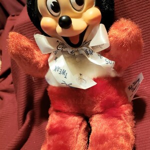 May include: A plush Mickey Mouse doll with a black head, large ears, and a smiling face. The doll has red arms, legs, and body, with white accents on the feet and a white bow tie. The bow tie has the word "Tweak" printed on it.