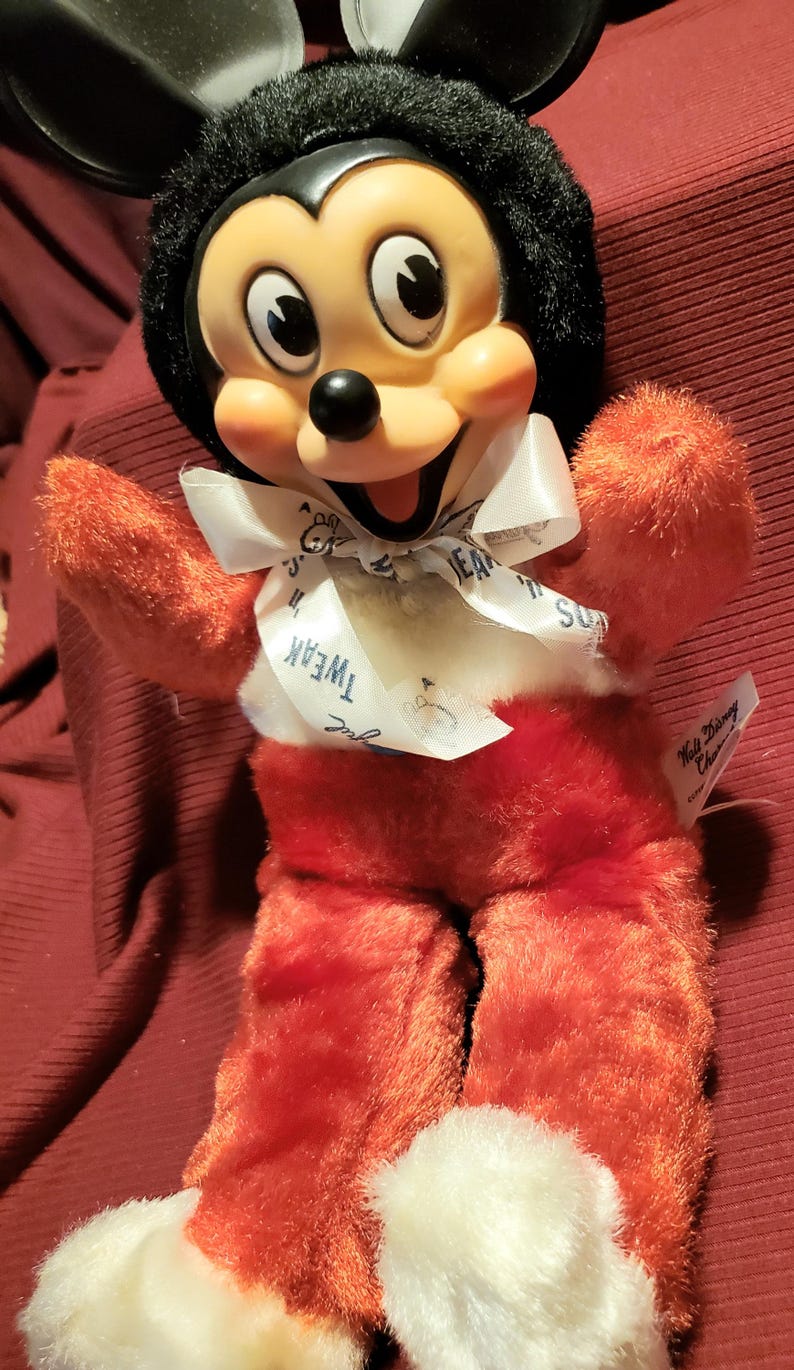 May include: A plush Mickey Mouse doll with a black head, large ears, and a smiling face. The doll has red arms, legs, and body, with white accents on the feet and a white bow tie. The bow tie has the word "Tweak" printed on it.