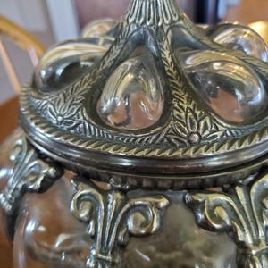 May include: Close-up of a decorative glass jar with a silver-colored lid and ornate detailing. The lid features a floral design and teardrop-shaped accents. The jar's body is clear, and the silver elements have an antique finish.