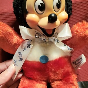 May include: Vintage Mickey Mouse plush toy with a black head, orange arms and legs, and red shorts. The toy has a white bow with the text "Tweak 'n Squeak". A blue button is on the belly. A Walt Disney Character tag is attached.