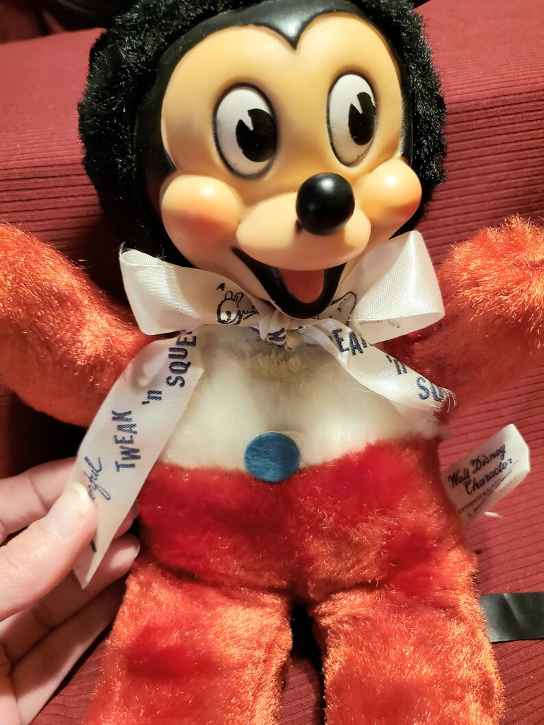 May include: Vintage Mickey Mouse plush toy with a black head, orange arms and legs, and red shorts. The toy has a white bow with the text "Tweak 'n Squeak". A blue button is on the belly. A Walt Disney Character tag is attached.