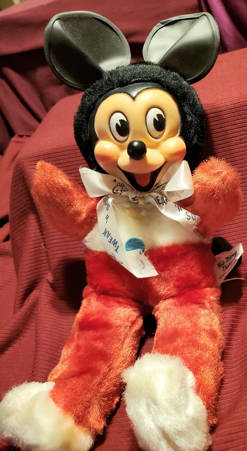 May include: A vintage Mickey Mouse plush toy. The toy features a black head with large ears, a smiling face, and red plush body with white accents. A white ribbon with text is around the neck. The toy is a collectible item.