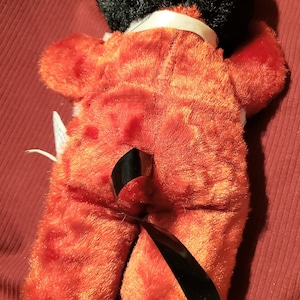 May include: A plush toy in the shape of a fox, viewed from the back. The fox has orange fur with white accents on the feet and a black head. It has a black, shiny, curved piece attached to its back.