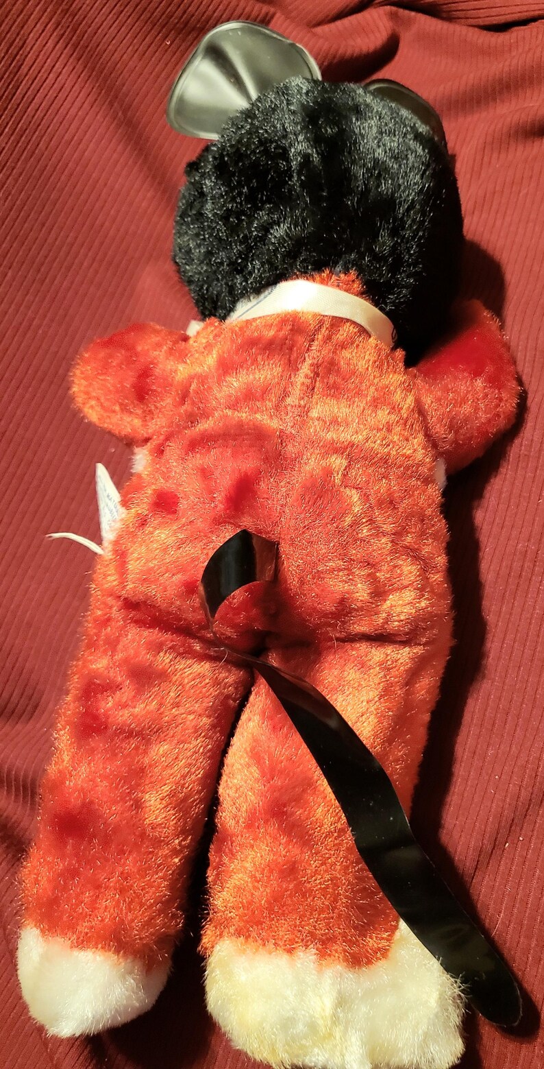 May include: A plush toy in the shape of a fox, viewed from the back. The fox has orange fur with white accents on the feet and a black head. It has a black, shiny, curved piece attached to its back.