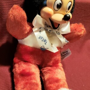 May include: Vintage Mickey Mouse plush toy. The plush has a black head with large ears, a yellow face, and a white bow with the text "Tweak". The body is red with white accents and white fluffy feet. A Disney Character tag is attached.