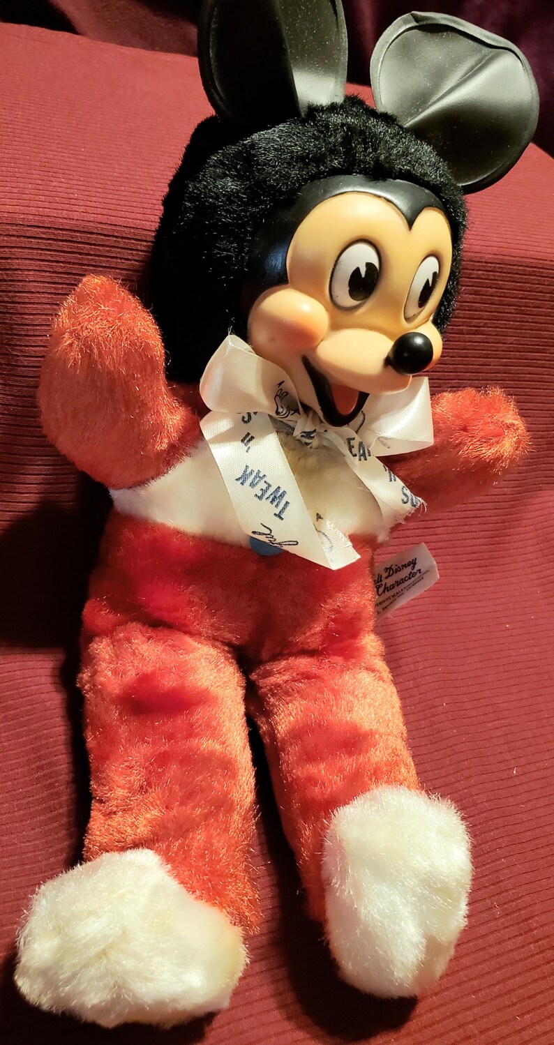 May include: Vintage Mickey Mouse plush toy. The plush has a black head with large ears, a yellow face, and a white bow with the text "Tweak". The body is red with white accents and white fluffy feet. A Disney Character tag is attached.