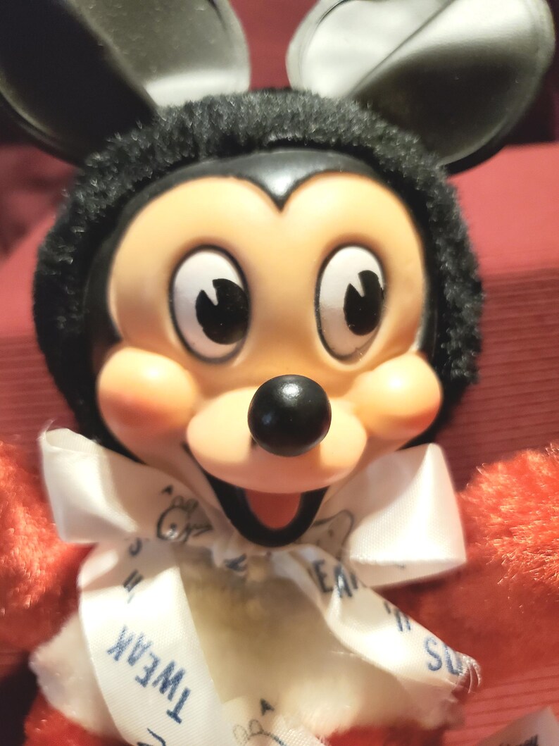 May include: A plush Mickey Mouse doll with a black head and ears, a tan face, and a black nose. The doll has a white bow tie with the word "Tweak" visible. The doll's arms are red.