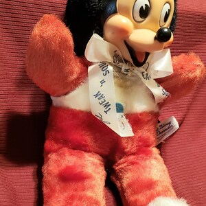 May include: Vintage Mickey Mouse plush doll with a black head and ears, a cream-colored face, and red body and legs. The doll has a white ribbon around its neck with the text "Tweak 'n' Squeak".