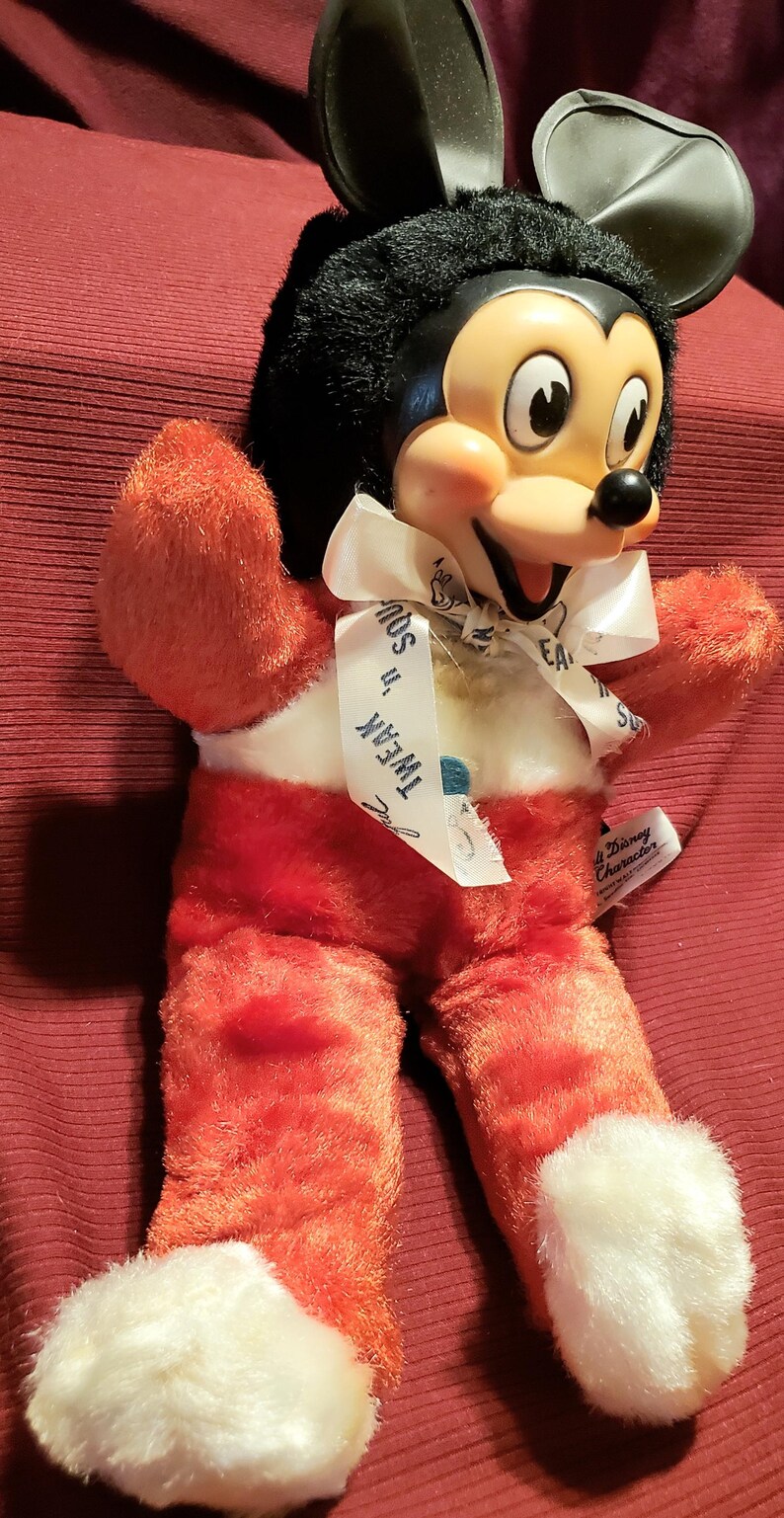 May include: Vintage Mickey Mouse plush doll with a black head and ears, a cream-colored face, and red body and legs. The doll has a white ribbon around its neck with the text "Tweak 'n' Squeak".