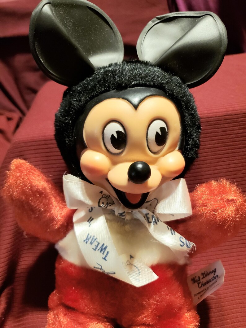May include: A vintage Mickey Mouse plush toy with black ears and a black fur head covering. The plush has a white ribbon with blue text and red arms and body. A Walt Disney Character tag is visible.