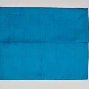 May include: A rectangular envelope-style clutch bag in a vibrant turquoise blue color. The exterior features a subtle, textured, slubbed fabric finish with a horizontal flap that folds over the main body, creating a clean and simple silhouette.