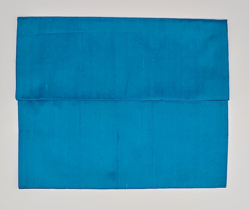 May include: A rectangular envelope-style clutch bag in a vibrant turquoise blue color. The exterior features a subtle, textured, slubbed fabric finish with a horizontal flap that folds over the main body, creating a clean and simple silhouette.