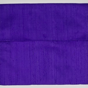 May include: A square, deep purple fabric item featuring a horizontal fold across the middle. The material displays a subtle, textured, slubbed appearance with vertical variations in the weave, creating a consistent, rich violet color throughout the surface.