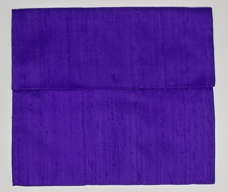 May include: A square, deep purple fabric item featuring a horizontal fold across the middle. The material displays a subtle, textured, slubbed appearance with vertical variations in the weave, creating a consistent, rich violet color throughout the surface.