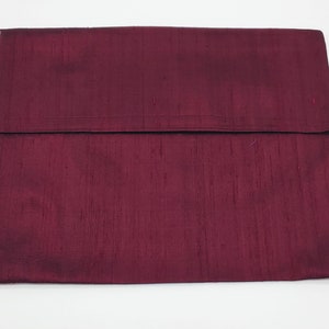 May include: A rectangular clutch bag in a deep burgundy color with a visible slubbed texture throughout the fabric. The bag features a large, wide flap that folds over the main body, creating a clean, horizontal line across the center.