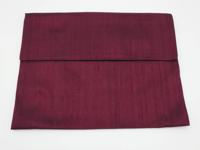 May include: A rectangular clutch bag in a deep burgundy color with a visible slubbed texture throughout the fabric. The bag features a large, wide flap that folds over the main body, creating a clean, horizontal line across the center.