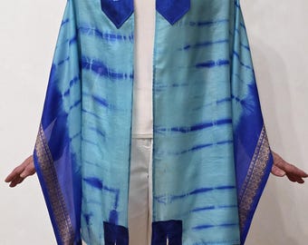 May include: A blue and turquoise prayer shawl with dark blue accents and gold trim. The shawl has a tie-dye pattern and features embroidered Hebrew text on the collar. The corners have tassels.