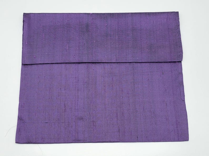 May include: A rectangular purple pillow cover featuring a visible slubbed texture throughout the fabric. The cover has a horizontal fold line across the upper section, creating a flap-like appearance with a consistent deep violet color tone.