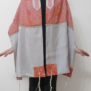 May include: A pink and white prayer shawl with embroidered floral designs and silver Hebrew text on the tallit straps. The shawl has a white satin lining and white fringes.