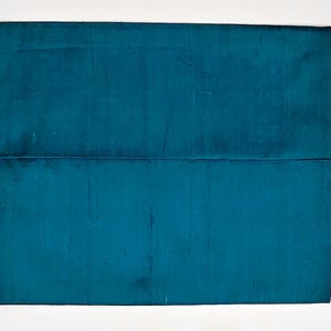 May include: A rectangular teal clutch bag with a smooth, slightly iridescent fabric finish. The bag features a horizontal flap closure that extends across the width of the item, creating a clean, structured silhouette with a consistent deep blue-green color.
