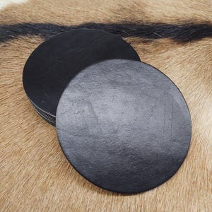 May include: A stack of black, round coasters made of a textured material. The coasters are displayed on a brown and tan patterned surface.