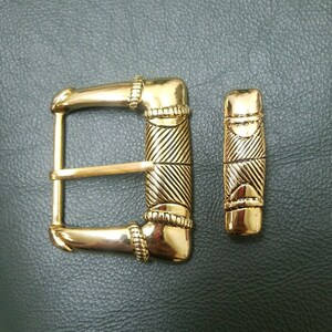 May include: A gold-colored belt buckle and matching keeper. The buckle has a rectangular frame with a diagonal striped design on the center bar. The keeper is a smaller, rectangular piece with a similar design. Both pieces have decorative details.