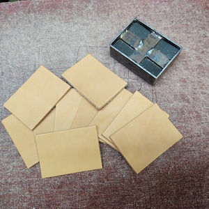 Puede incluir: Primer plano de una pila de seis cuadrados de cuero marrón claro, con una herramienta de metal con un diseño en forma de cruz en el fondo.