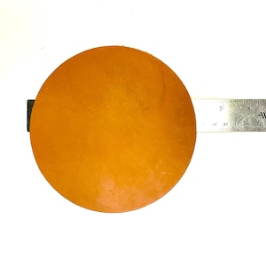 May include: A round, orange-brown leather craft disc, approximately 7 inches in diameter, with a ruler in the background.