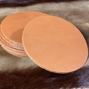 May include: A stack of circular, tan-colored leather coasters with smooth, flat surfaces and visible, raw-edged sides. The coasters are uniform in shape and size, featuring a natural, matte finish with subtle variations in the light brown leather grain.