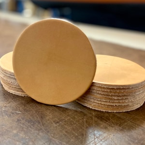 May include: A stack of round, tan leather coasters. The top coaster is larger than the others and is laying flat on a wooden surface.