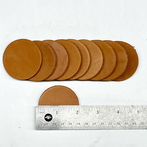 May include: A collection of tan leather circles, arranged in a row, with one cut in half. A ruler shows the diameter of the circles is approximately 2 inches. The leather has a natural, textured appearance.
