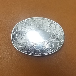 Puede incluir: Hebilla de cinturón ovalada plateada con intrincados adornos florales. La hebilla tiene una superficie pulida y reflectante y un borde detallado. El diseño presenta un área central sin adornos, rodeada por el patrón ornamentado. Un accesorio Western clásico.