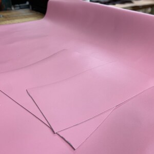 May include: Two pieces of pink leather fabric, one larger than the other, are laid out on a table. The fabric has a smooth, glossy finish.