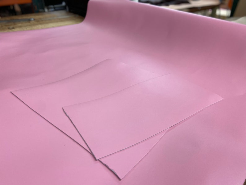 May include: Two pieces of pink leather fabric, one larger than the other, are laid out on a table. The fabric has a smooth, glossy finish.