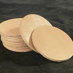 May include: A stack of light tan leather circles, possibly coasters, with a smooth surface. Several circles are stacked, with a few more lying on top. The leather has a natural, unfinished look.