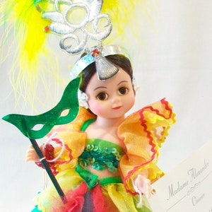 May include: A Madame Alexander Classic doll in a colorful costume. The doll wears a headdress with yellow and green feathers, a green mask, and a multi-layered dress in yellow, green, and red. The doll is holding a black stick with a red bead.