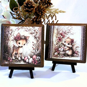 May include: Three square framed art pieces on small black easels. Each features a different woodland creature: a mouse, a deer, and a squirrel, all adorned with floral crowns. The artwork has a vintage, storybook aesthetic with soft colors.