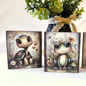 May include: Three decorative wooden blocks featuring whimsical illustrations of a turtle, a frog, and a snail. Each block has a distressed, vintage look with floral accents. A vase with greenery and a bow is in the background.