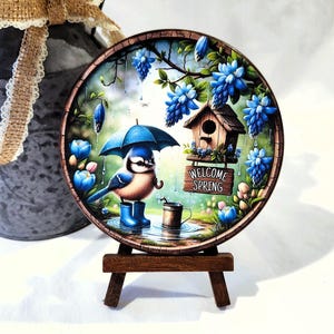 May include: A round decorative plaque featuring a blue bird under an umbrella, wearing blue boots, and standing in the rain. The plaque has a wooden birdhouse and the words "WELCOME SPRING". The plaque is displayed on a small wooden easel.