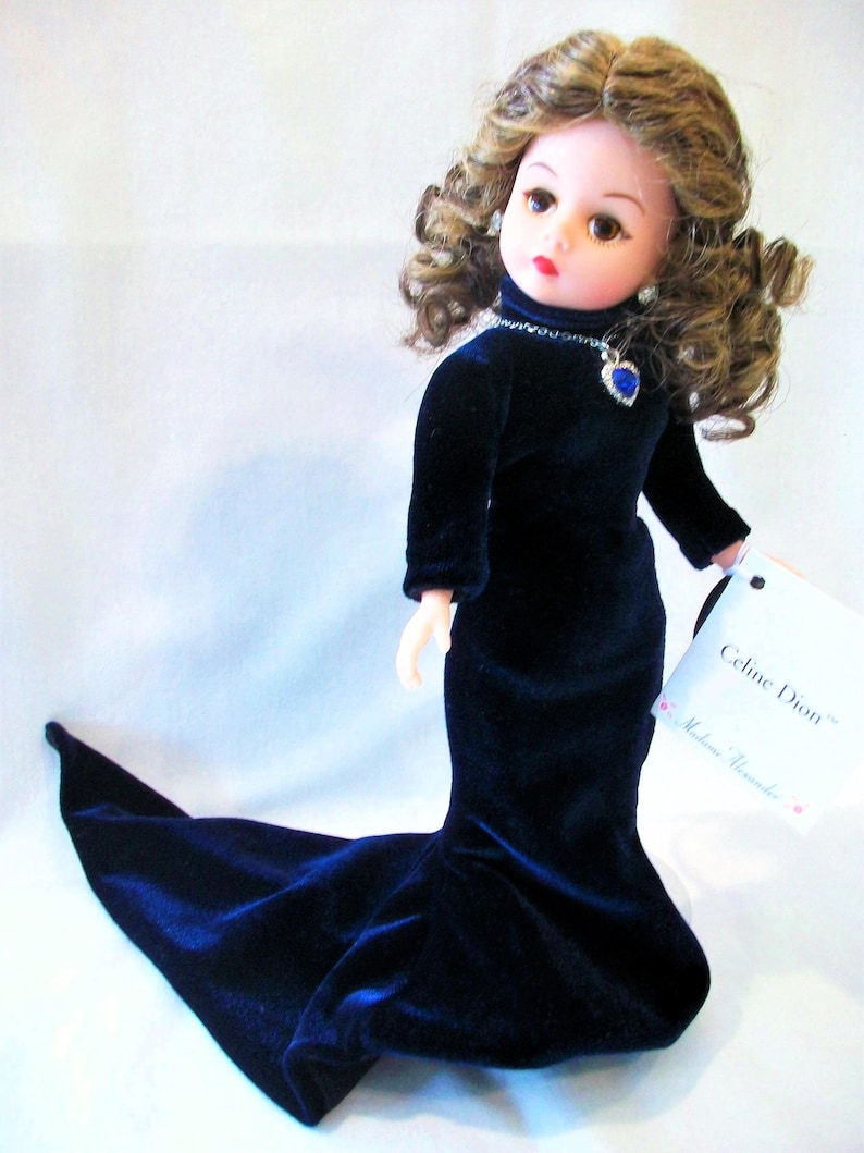 May include: A doll in a dark blue velvet gown with a long train. The doll has brown curly hair, red lips, and a blue heart-shaped pendant necklace. The tag reads "Celine Dion Madame Alexander".