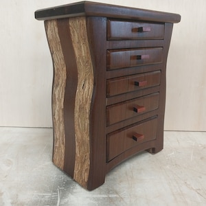 May include: A small wooden dresser with five drawers and a curved front. The dresser is made of dark wood with light wood accents. The drawers have small wooden knobs.