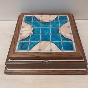May include: A wooden box with a decorative top featuring a blue and tan design. The top has a grid pattern with small pink dots. The box is made of dark brown wood with a lighter wood accent.