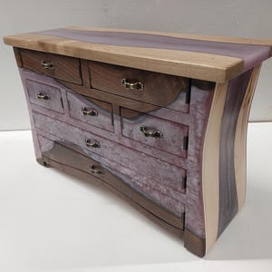 May include: A handcrafted wooden jewellery box with a unique design. The box features a light-coloured wood top with a purple resin inlay. The drawers and sides are a mix of dark and light wood with a purple and white marbled resin. Antique brass drawer pulls.