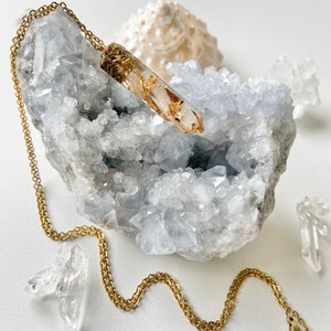 Pode incluir: Um colar de corrente dourada com um pingente de cristal de quartzo transparente que tem folhas de ouro embutidas. O pingente está apoiado em um leito de cristais brancos.