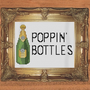May include: A framed cross-stitch design featuring a green and gold champagne bottle with the text "Poppin' Bottles" in black.