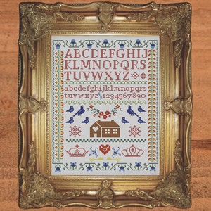 May include: A framed cross-stitch sampler with a white background and colorful embroidery. The sampler features the alphabet in uppercase and lowercase, numbers, birds, flowers, and a small house.