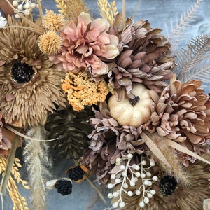 May include: A seasonal wreath with artificial flowers in autumnal tones of brown, beige, and peach. The design incorporates large chrysanthemum-style flowers, a small pumpkin, and decorative berries. Dried grasses and foliage provide texture and depth to the arrangement.