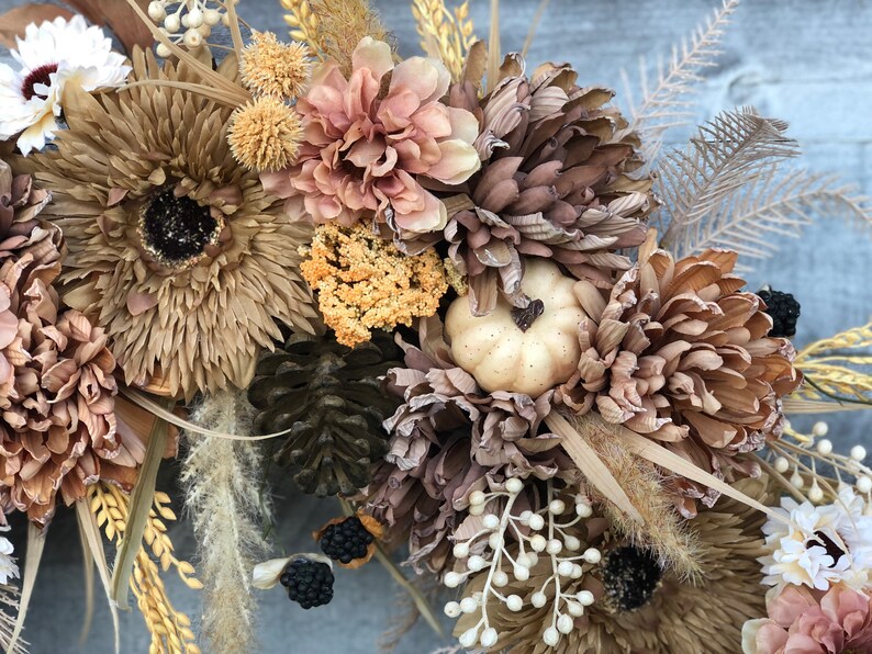 May include: A seasonal wreath with artificial flowers in autumnal tones of brown, beige, and peach. The design incorporates large chrysanthemum-style flowers, a small pumpkin, and decorative berries. Dried grasses and foliage provide texture and depth to the arrangement.
