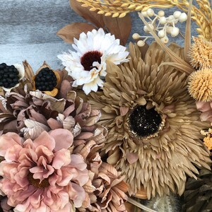 May include: Close-up of a decorative wreath with dried flowers and foliage in neutral tones. The wreath includes large, textured flowers in shades of brown, beige, and pink, with accents of white and black. The background is a weathered, grey wooden surface.