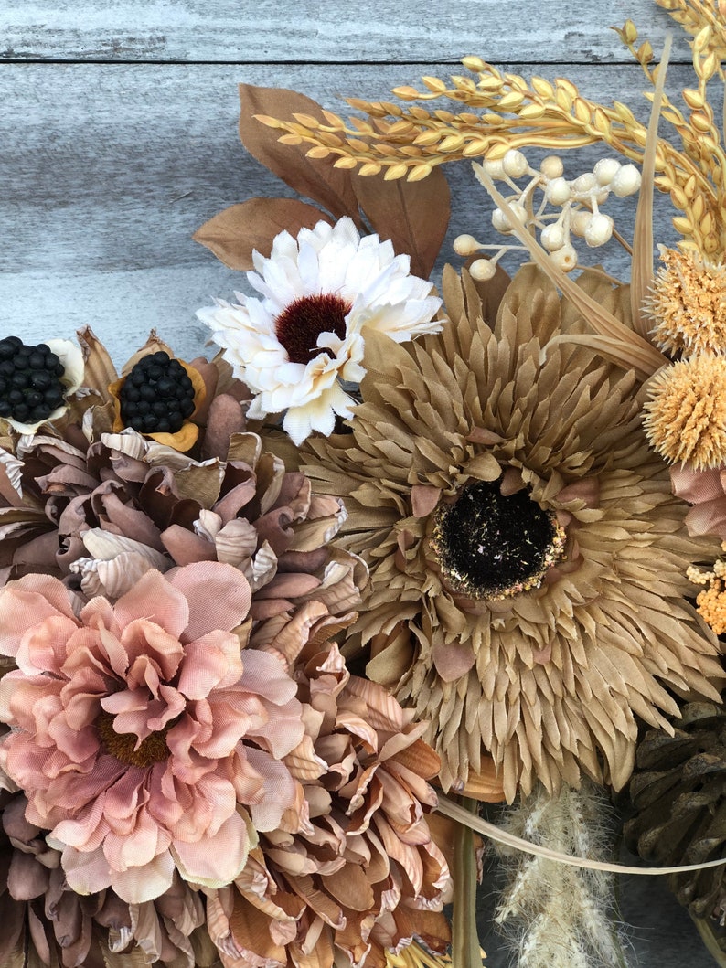 May include: Close-up of a decorative wreath with dried flowers and foliage in neutral tones. The wreath includes large, textured flowers in shades of brown, beige, and pink, with accents of white and black. The background is a weathered, grey wooden surface.